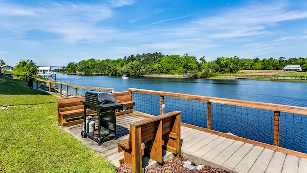 A wooden deck with a grill and seating area overlooking a river, surrounded by greenery and a clear sky. - Waterway views, beautiful sunsets, pools, hot tubs, balcony, grilling & beaches (North Myrtle Beach)