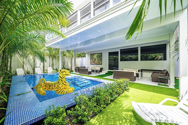 Zona de piscina exterior moderna amb una decoració de tigre realista, envoltada de vegetació exuberant i una zona de seients coberta amb mobiliari d'exterior.