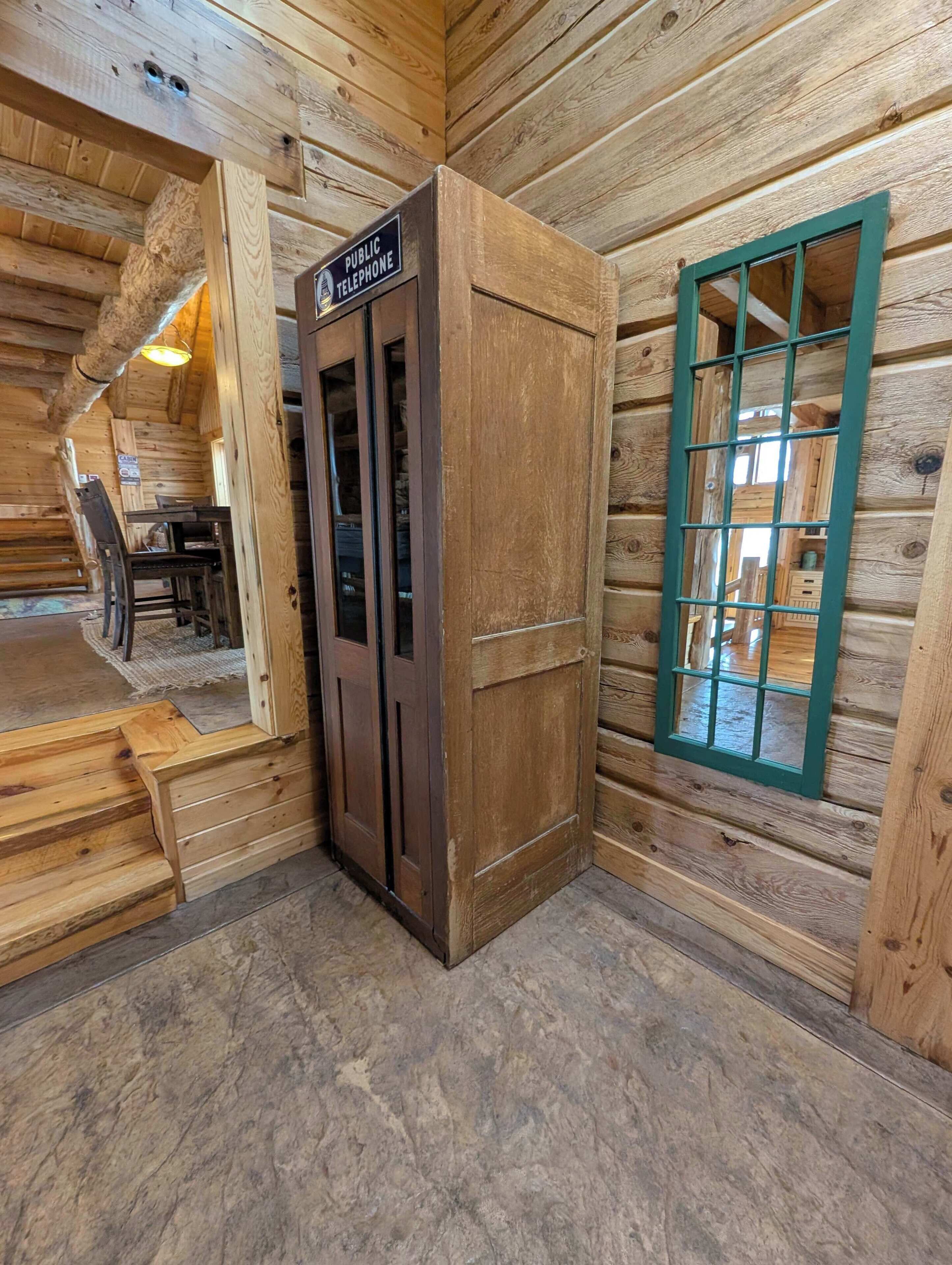 L’intérieur d’une cabane en bois, avec un téléphone public, une fenêtre à cadre vert et un escalier menant à un niveau supérieur.