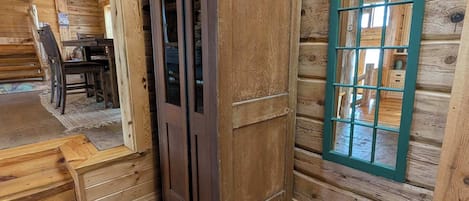 Interior de una cabaña de madera con un teléfono público, una ventana de marco verde y una escalera que sube a un nivel superior.