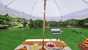 A wooden picnic table with a white umbrella, a grill, and a bowl of strawberries.