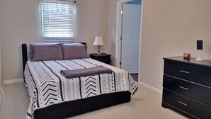 Een slaapkamer met een bed, nachtkastje, kapstok, plafondventilator, raam met jaloezieën en een deur.
