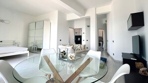 Una habitación moderna y bien iluminada con una mesa de vidrio, muebles blancos, una cama, un sofá y un televisor.