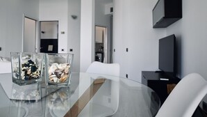 Un comedor moderno con una mesa de vidrio, dos jarrones de vidrio con conchas marinas, sillas blancas y un televisor de pantalla plana montado en la pared.