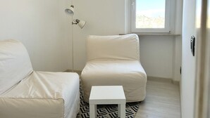 Una habitación pequeña y blanca con una cama, una silla y una mesita. Hay una ventana con vistas a edificios.