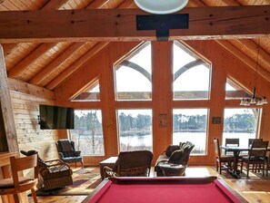 Una cabaña acogedora con mesa de billar, muebles de madera y grandes ventanales con vistas a un lago.