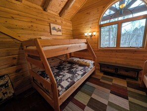 A bunk bed in a cabin with a checkered rug, a window with a view of trees, and a wall-mounted light fixture.
