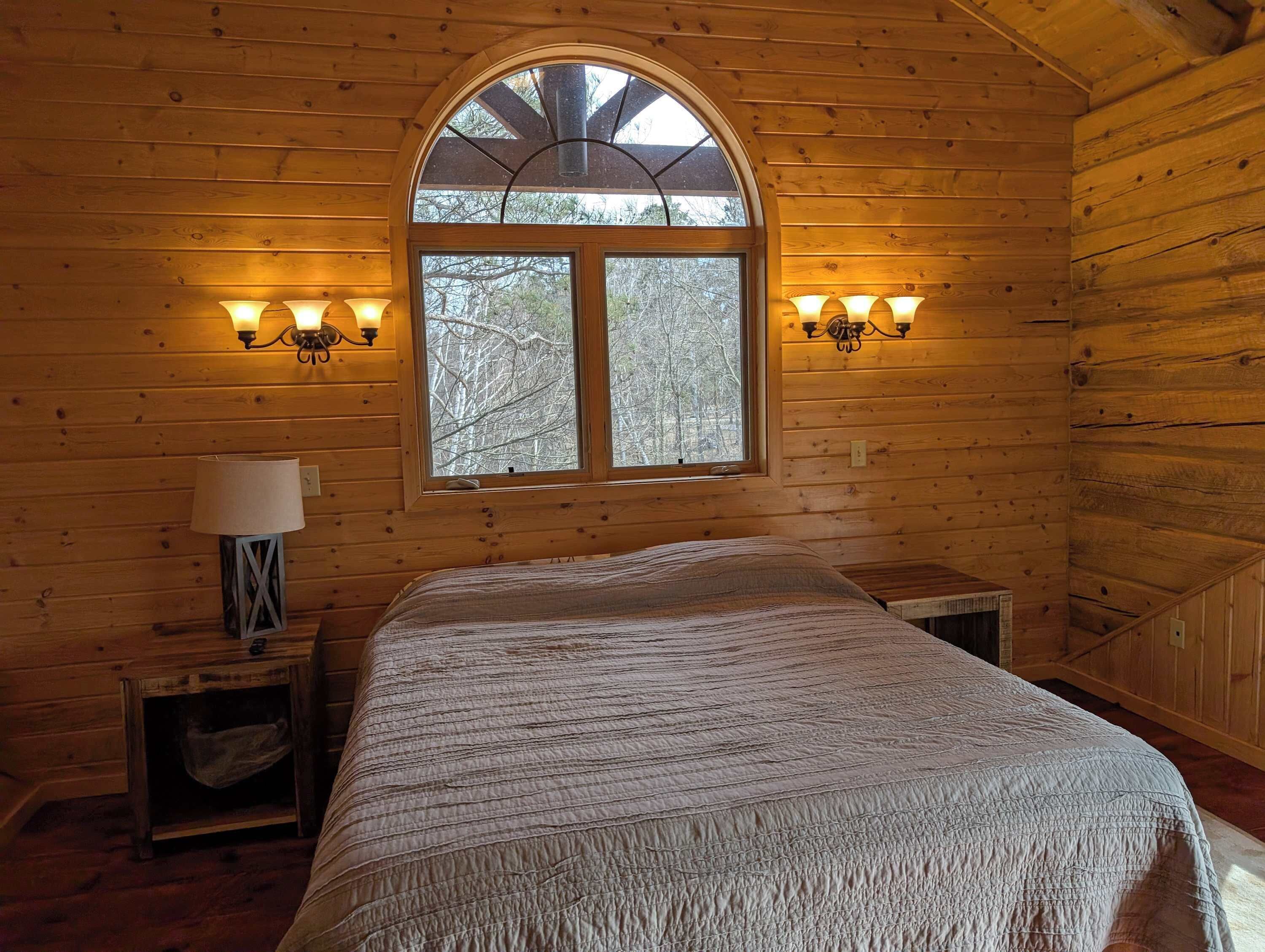 A wooden cabin bedroom with a large bed, two bedside lamps, a window with a view of trees, and a lamp on a nightstand.