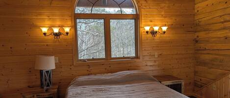 A wooden cabin bedroom with a large bed, two bedside lamps, a window with a view of trees, and a lamp on a nightstand.