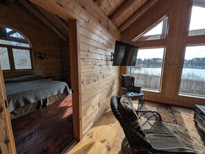 Interior de una cabaña con cama, televisor, sillón y vista al lago.