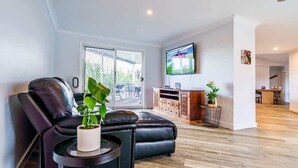 A modern living room with a black leather sofa, a small round table with a plant, a flat-screen TV mounted on the wall, and a sliding glass door leading to an outdoor area. - Mudgeeraba family home with hot tub & garden (Mudgeeraba)
