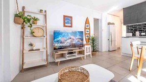 A modern living room with a flat-screen TV, a wooden shelf with plants, a surfboard decoration, and a kitchen area in the background.