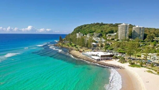 Ein Küstenresort mit Strandgrundstücken, einem Sandstrand und einer Kulisse aus üppiger Vegetation und hohen Gebäuden.