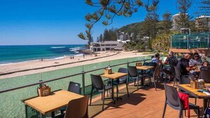 Ein Restaurant direkt am Strand mit Terrassenplätzen und Blick auf den Ozean, einem Pool und einem Gebäude im Hintergrund.