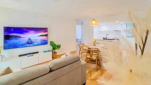 Ein modernes Wohnzimmer mit einer Couch, einer Essecke mit Stühlen und einem Tisch, einer Küche mit Spüle und Herd sowie einem an der Wand montierten Fernseher.