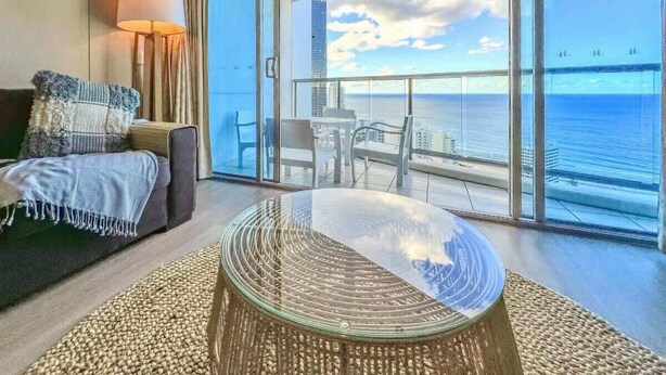 Uma sala de estar moderna com uma mesa de vidro, um sofá com manta e vista para o oceano através de grandes janelas.