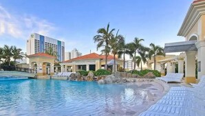 Área de piscina em resort com espreguiçadeiras, palmeiras e um edifício com telhado vermelho.