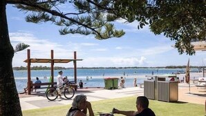 Un espace aménagé au bord de la plage, avec une pergola en bois, un vélo et des personnes se détendant près de l’eau.