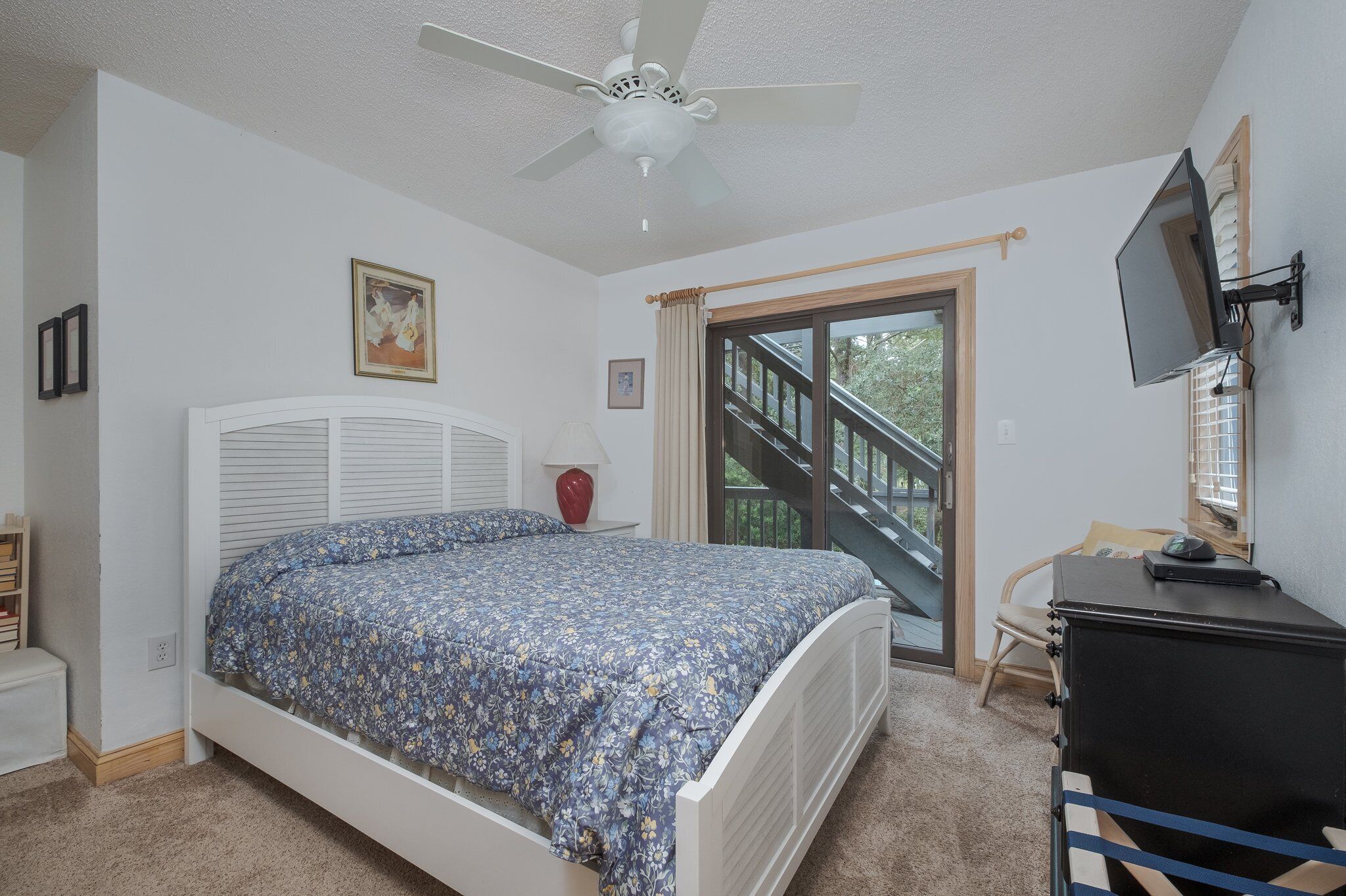 Une chambre à coucher comprenant un lit, un ventilateur de plafond, une télévision, un bureau et une fenêtre donnant sur un escalier.