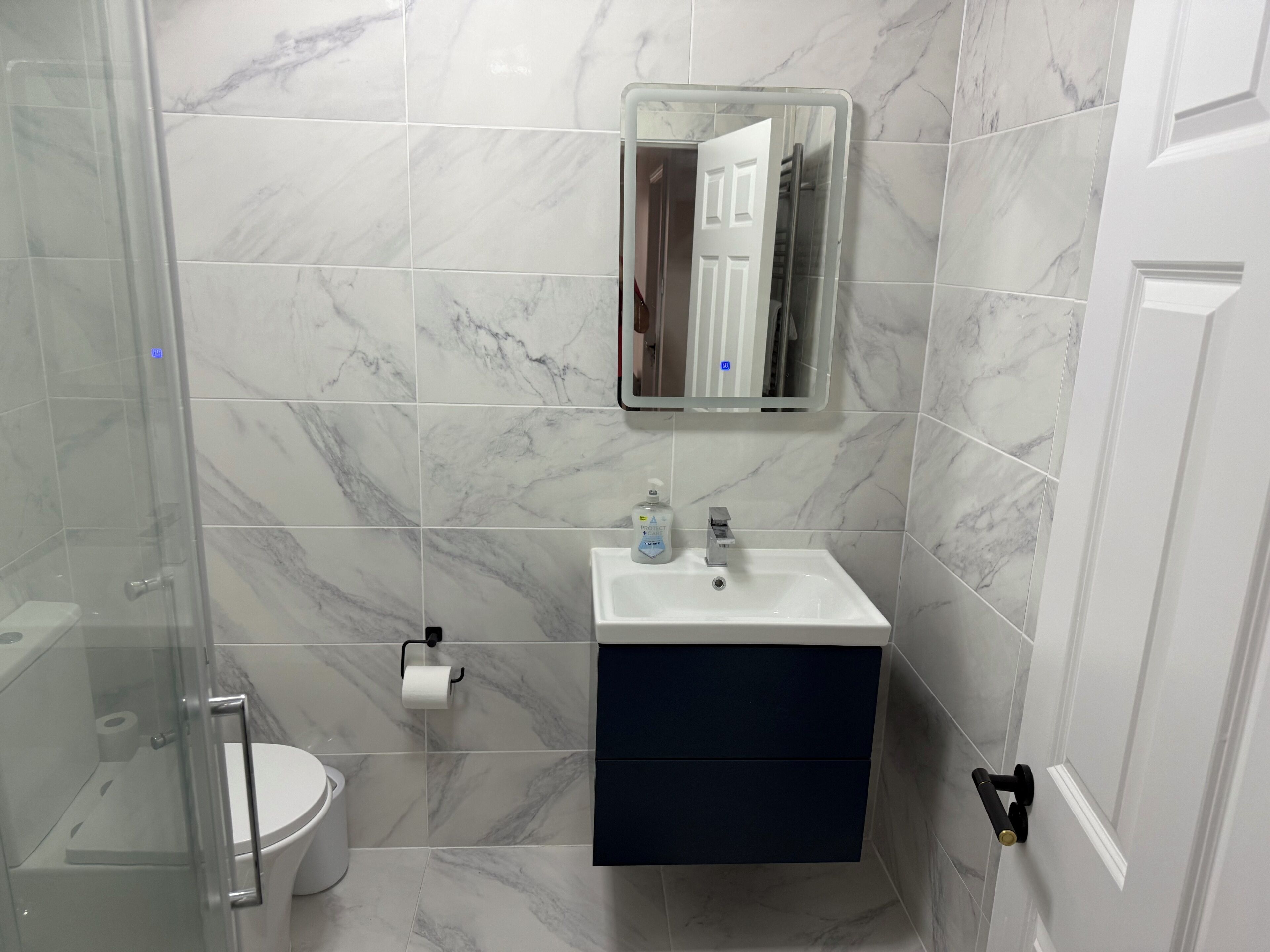 Un bany amb una cabina de dutxa de vidre, un vàter blanc, un lavabo amb un mirall a sobre i un armari fosc a sota.