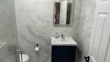 Un bany amb una cabina de dutxa de vidre, un vàter blanc, un lavabo amb un mirall a sobre i un armari fosc a sota.