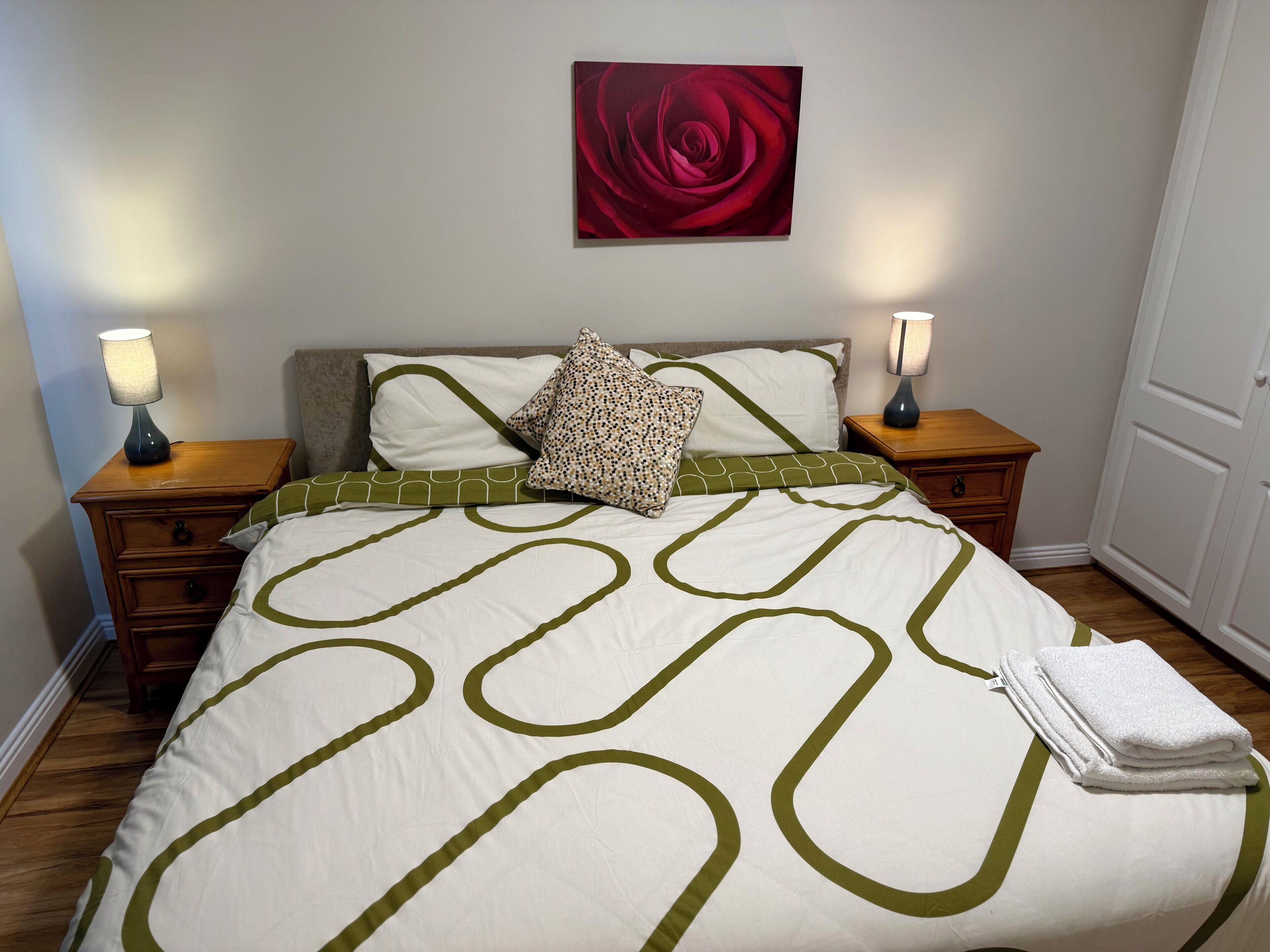 Ein ordentlich bezogenes Bett mit grün-weißem geometrischen Muster, zwei Nachttischlampen, ein Gemälde mit einer roten Rose an der Wand und eine Tür rechts.