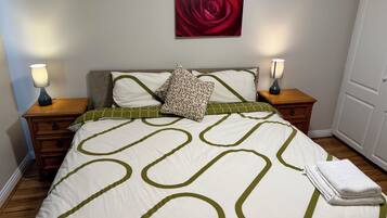 Ein ordentlich bezogenes Bett mit grün-weißem geometrischen Muster, zwei Nachttischlampen, ein Gemälde mit einer roten Rose an der Wand und eine Tür rechts.