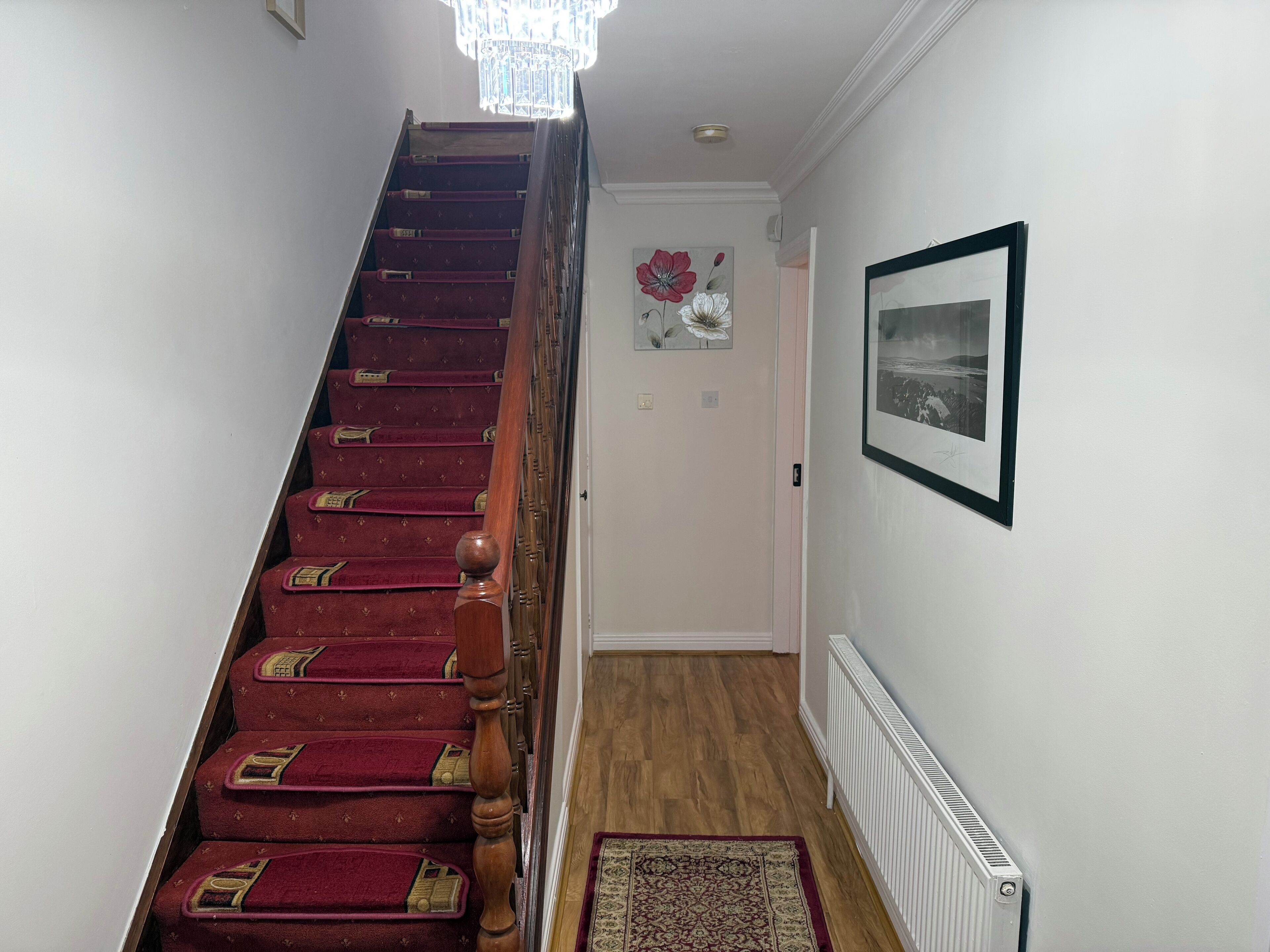 Eine Treppe mit roten Teppichstufen und einem hölzernen Geländer, die zu einem Raum mit Heizkörper und einem gerahmten Bild führt.