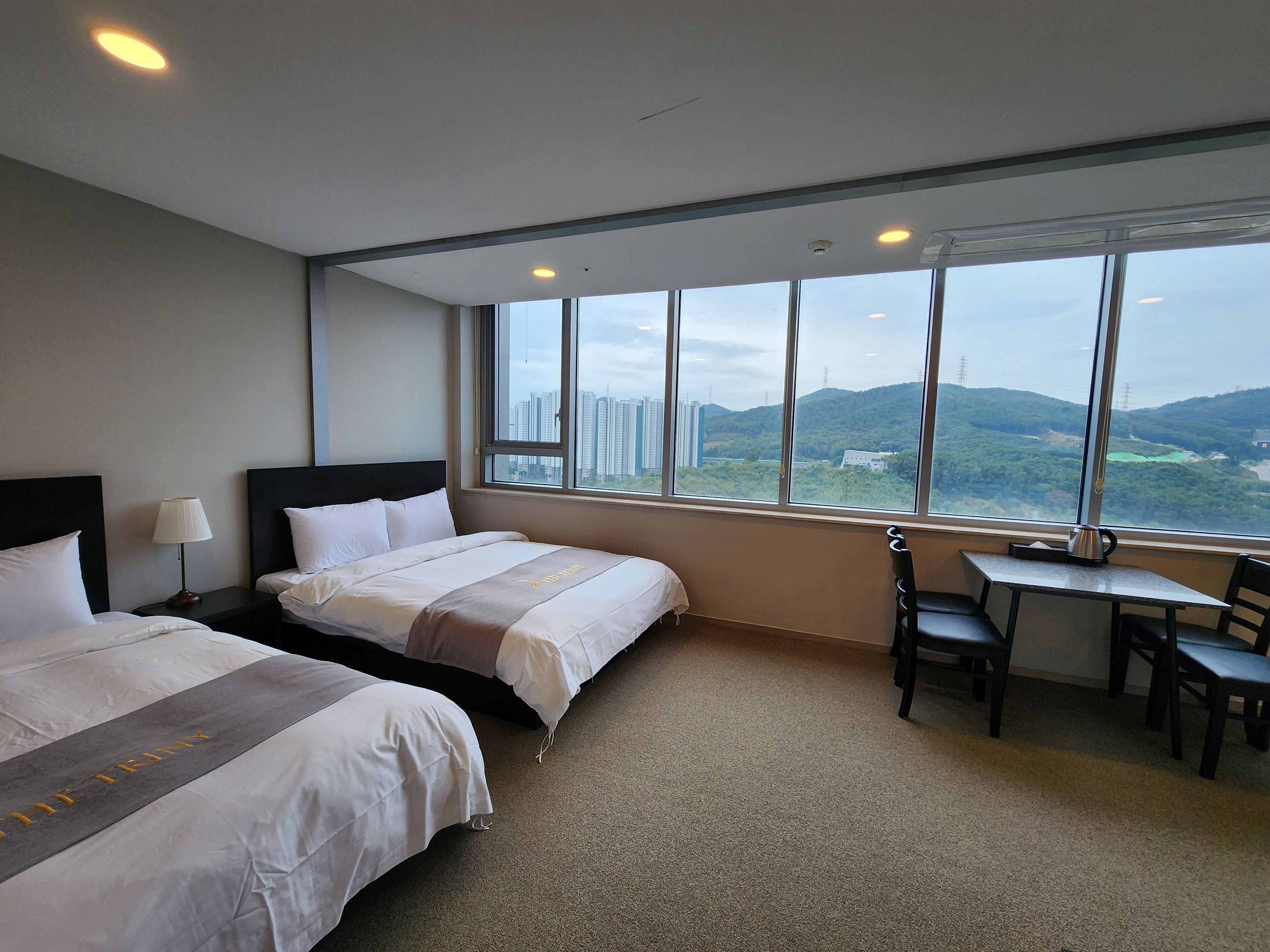 A hotel room with two beds, a desk, and a view of a cityscape through large windows.