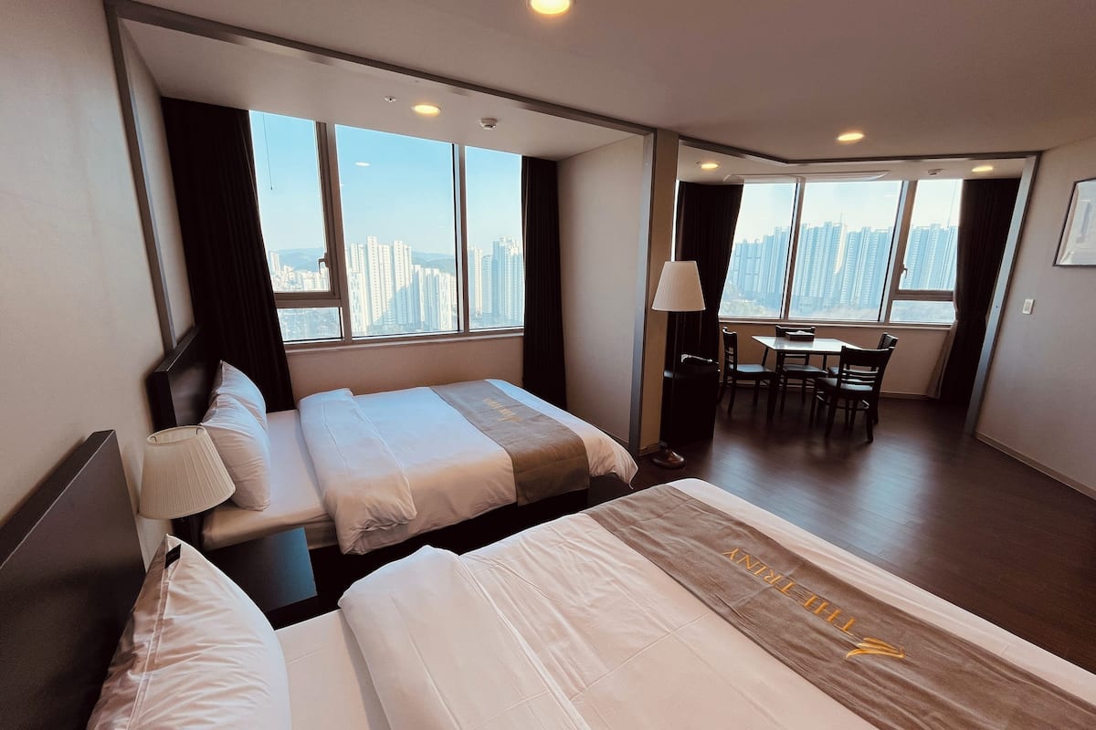 A hotel room with two beds, a dining table, and large windows offering a city view.