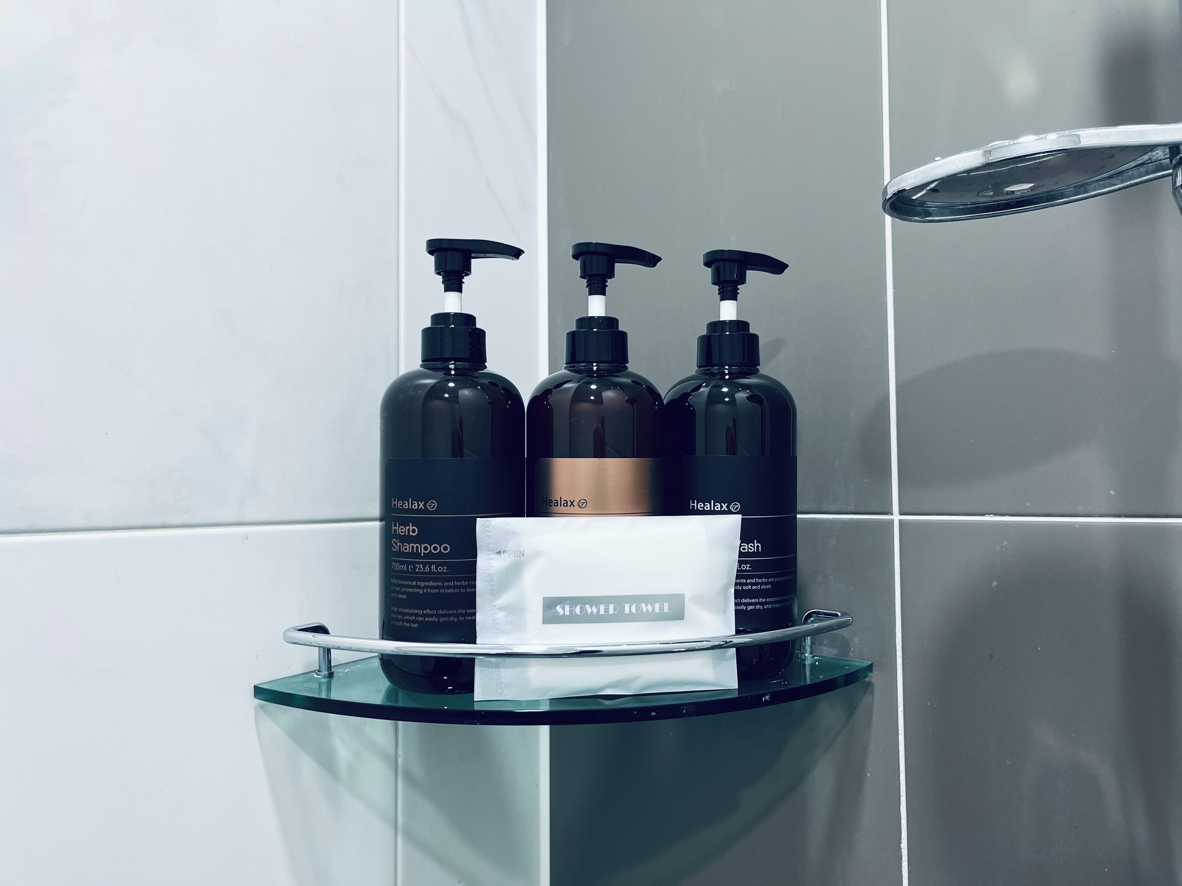A bathroom with a glass shelf holding three black bottles of Healax shampoo and conditioner, and a white towel.