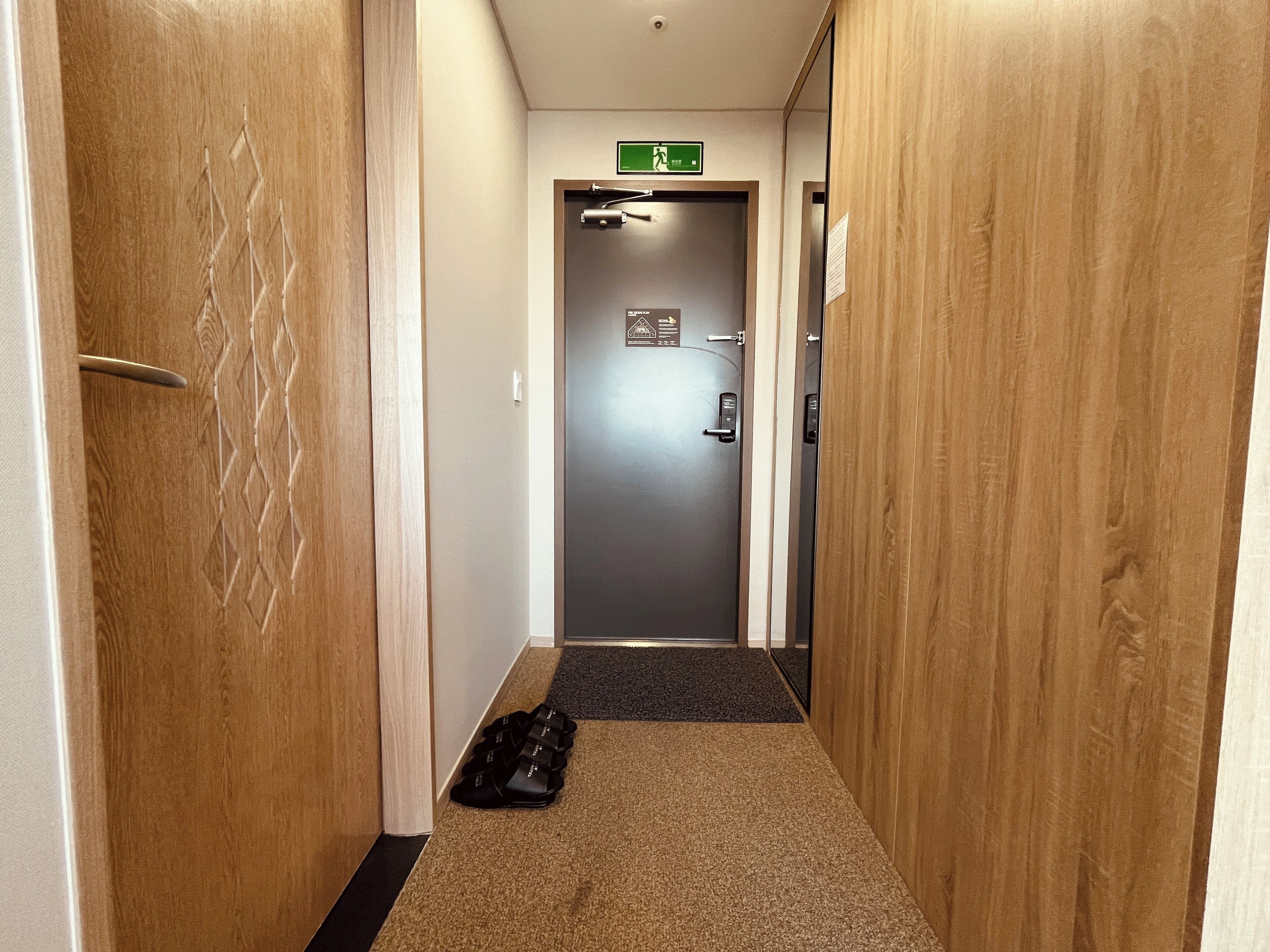 Un couloir d’hôtel avec des portes en bois, un sol recouvert de moquette et une porte sur laquelle est apposée une signalétique verte indiquant la sortie de secours.