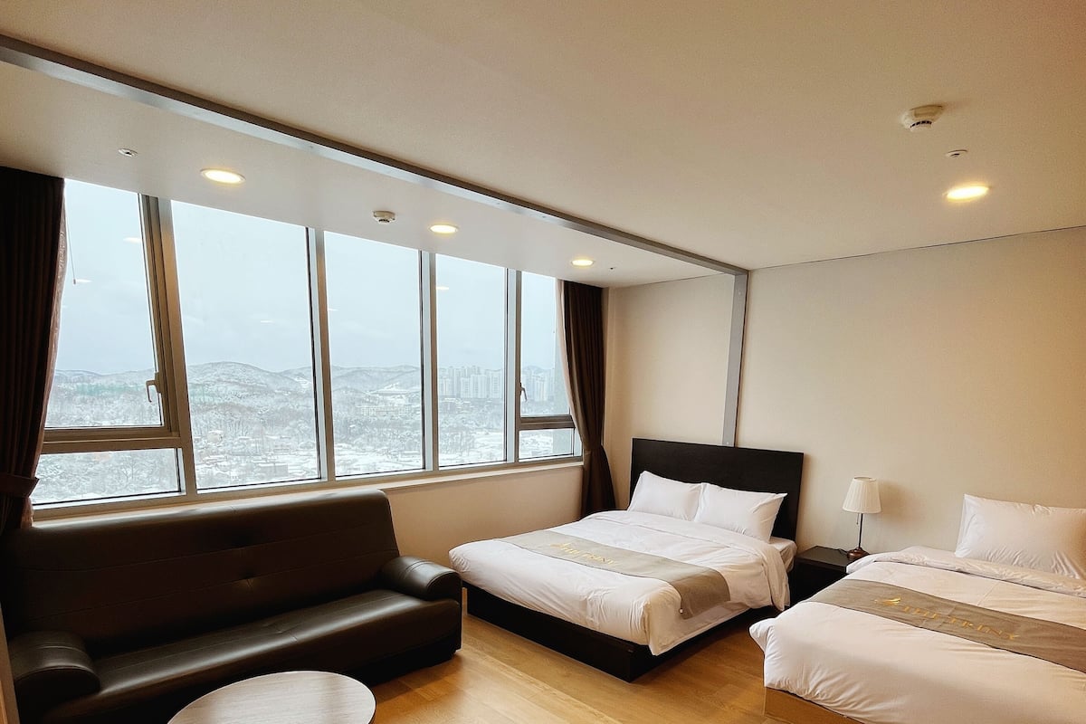 A hotel room with a large window, a sofa, two beds, and a small table.