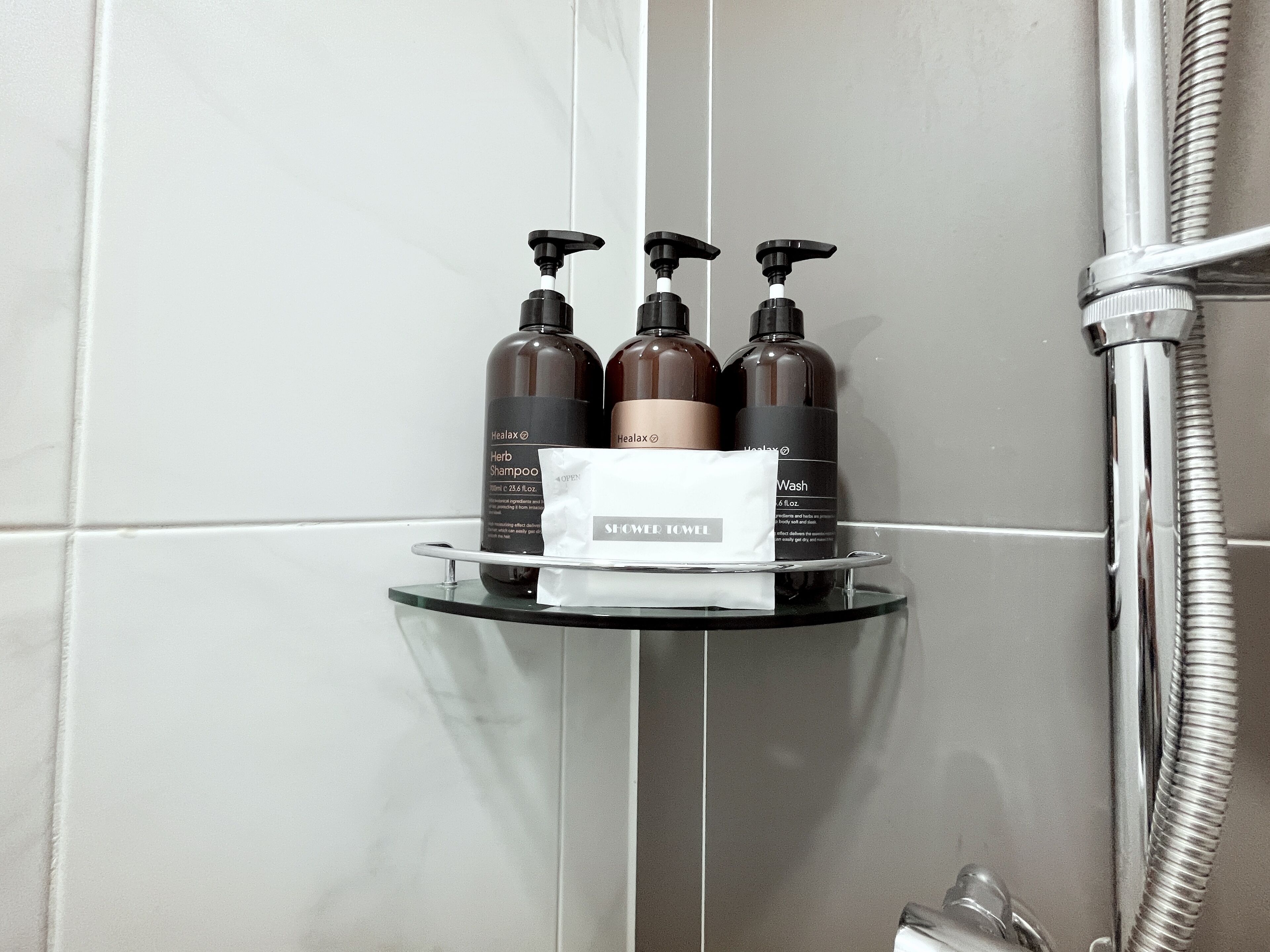 A bathroom with a glass shelf holding three bottles of shampoo and a shower towel.
