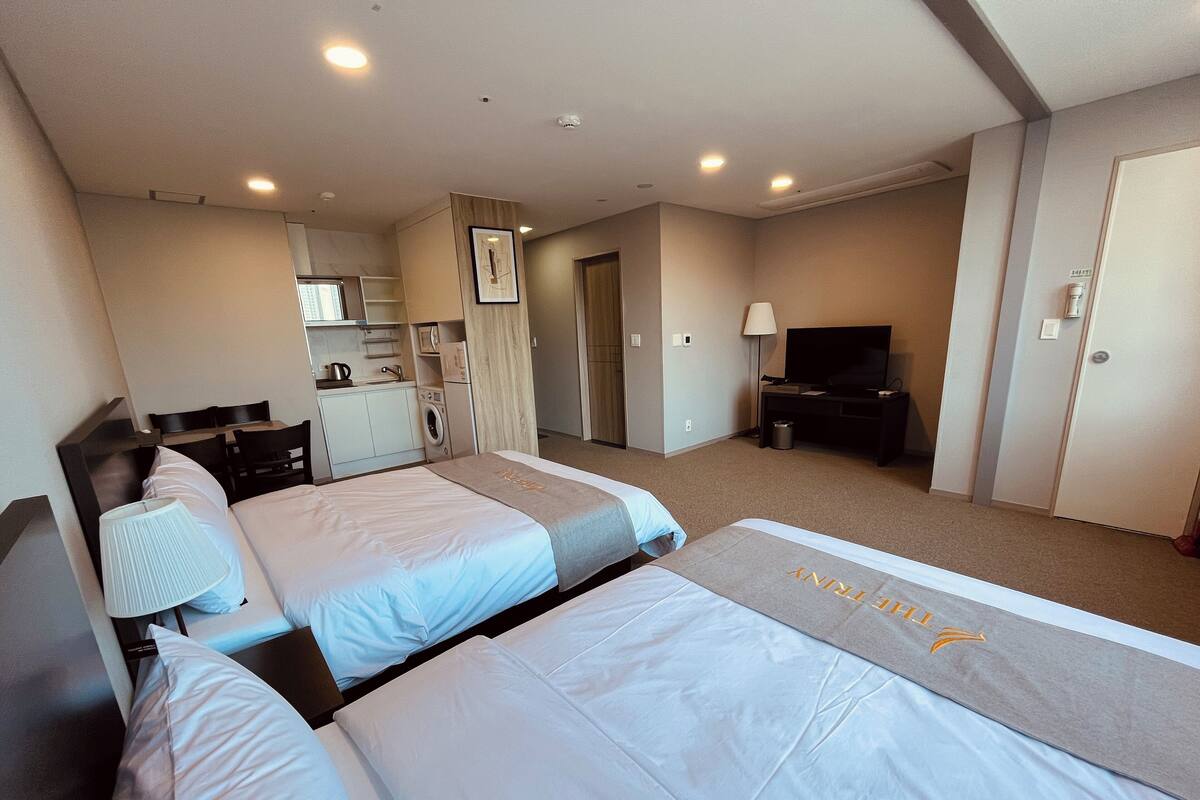 A hotel room with two beds, a desk, a TV, a washing machine, and a kitchenette.