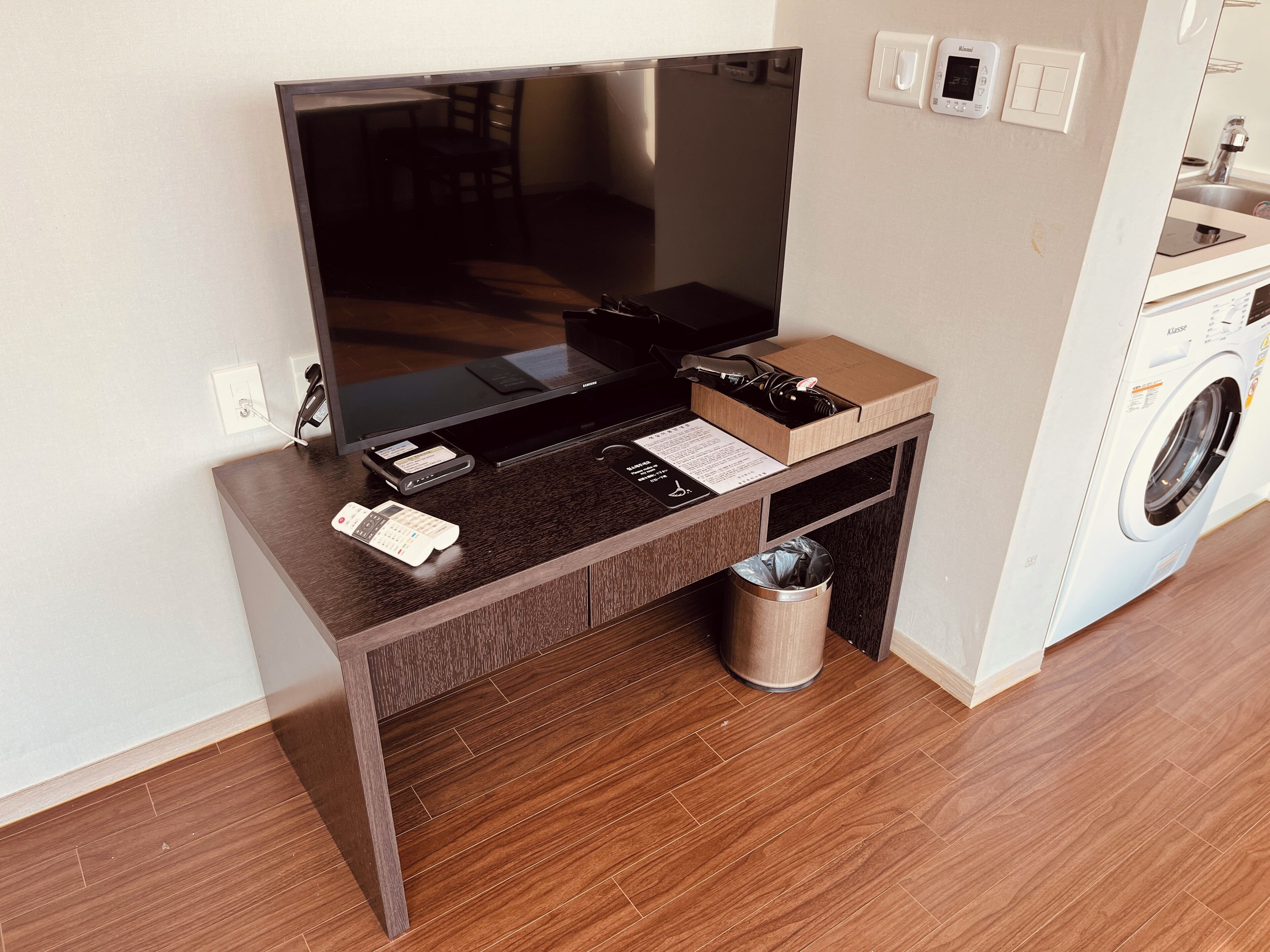 A modern living room with a flat-screen TV, a coffee table, a small trash bin, and a washing machine in the background.
