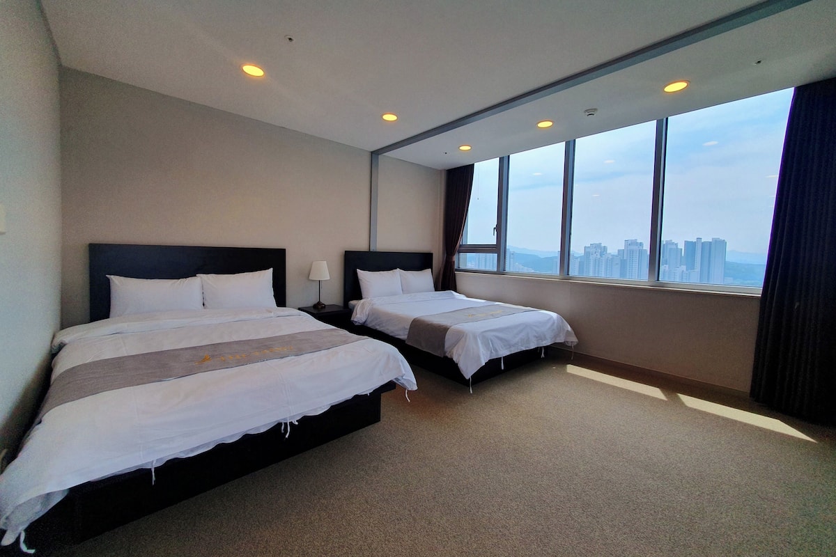 A hotel room with two beds, a large window with a city view, and a carpeted floor.