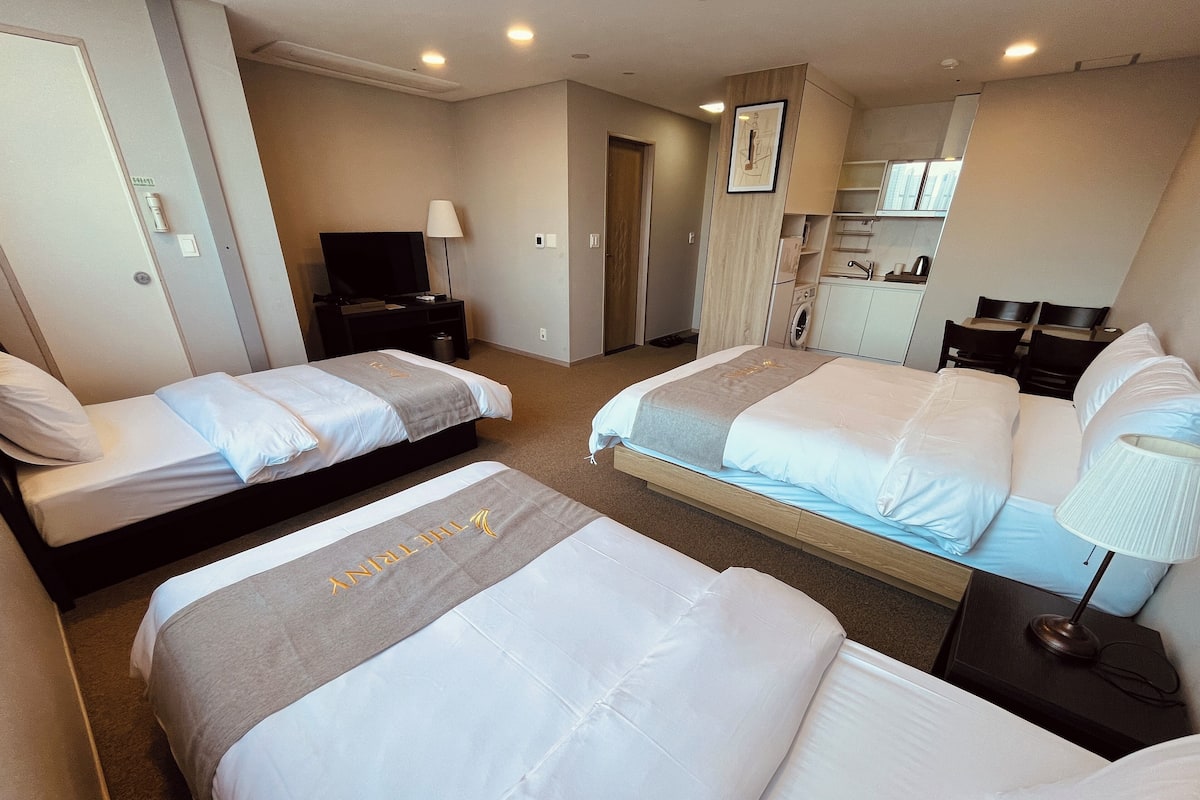 A hotel room with two beds, a TV, a desk, and a kitchenette.