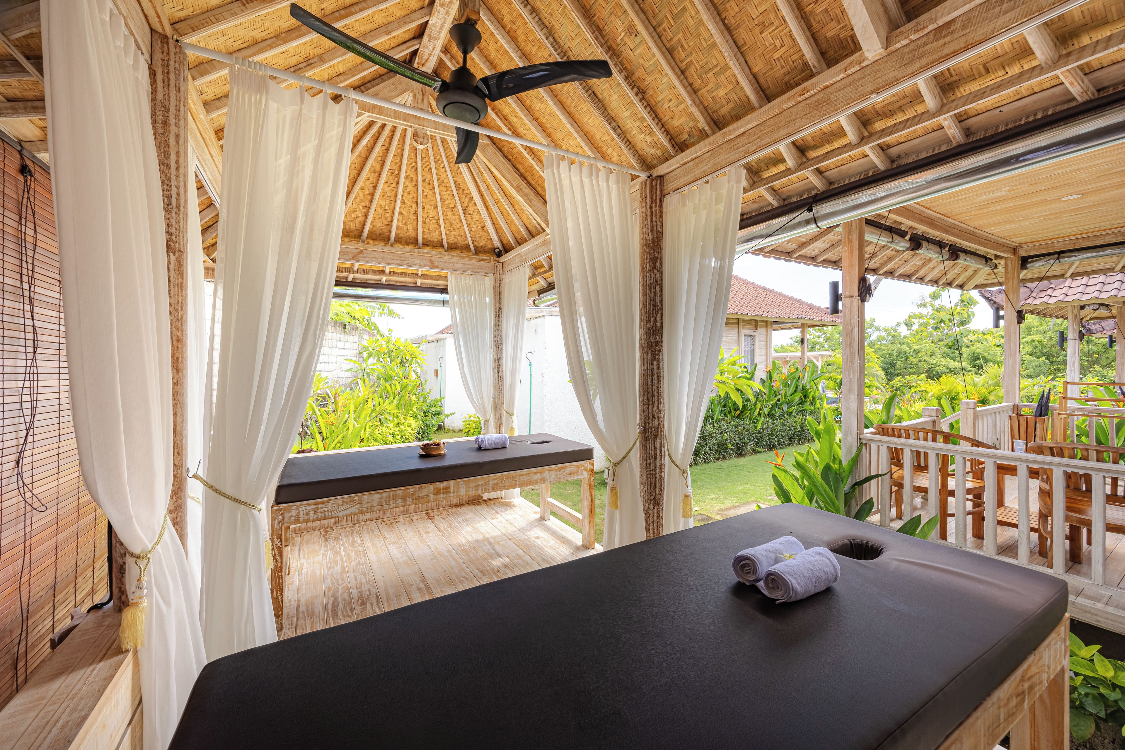 A massage table with a ceiling fan and white curtains in a wooden room with a view of greenery.