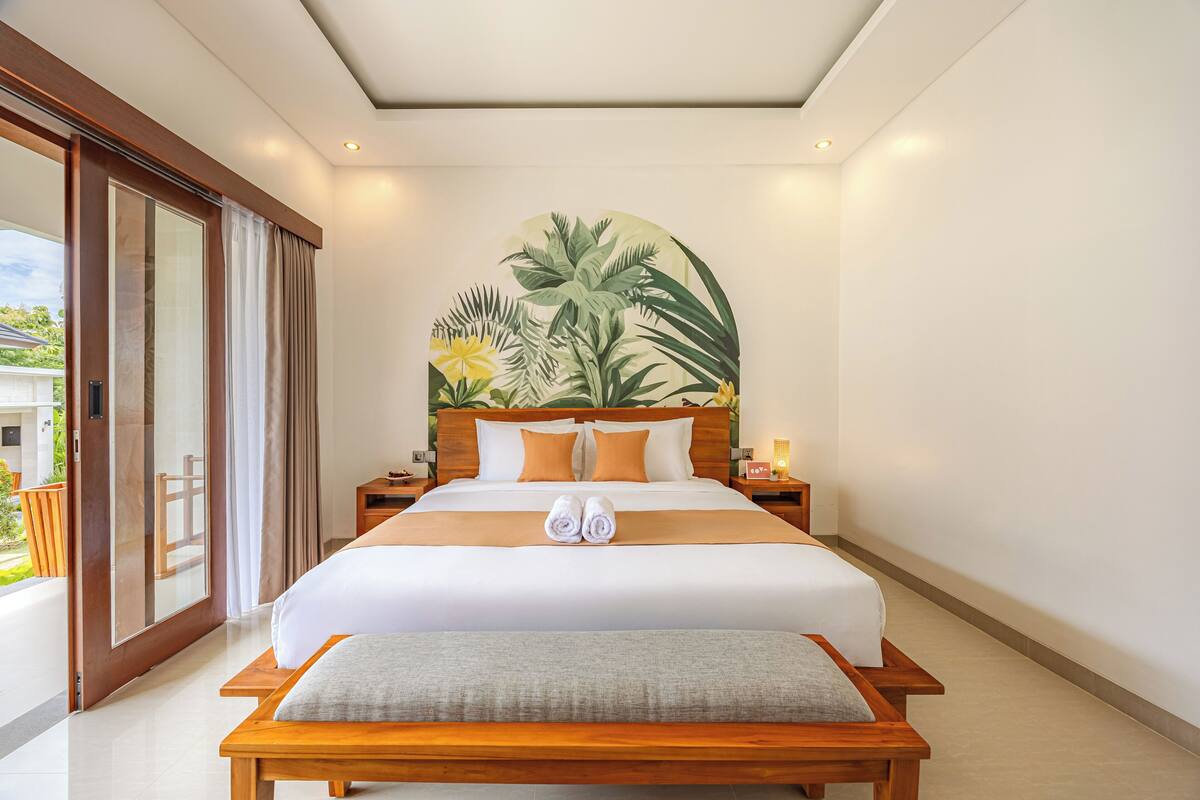 Una cama grande con cabecera de madera, un banco al pie de la cama y un cuadro de follaje tropical en la pared.