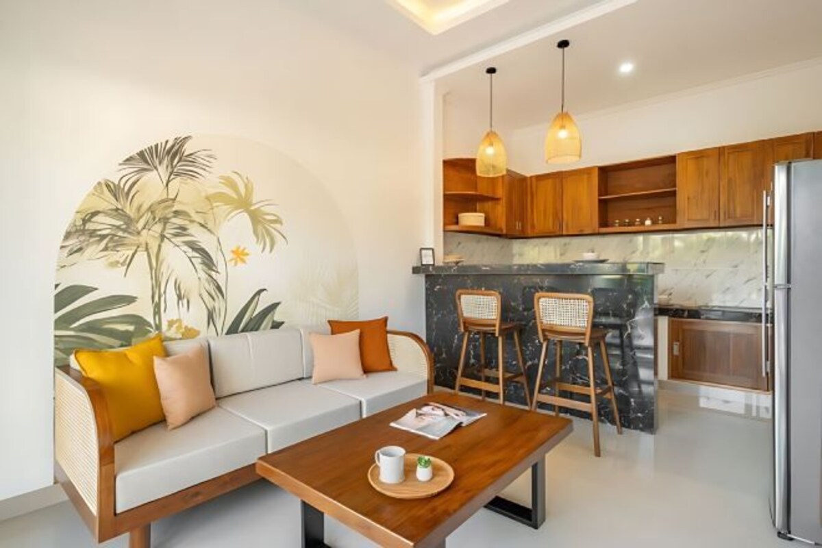 A modern living room with a sofa, coffee table, and bar stools. A kitchen with wooden cabinets and a refrigerator is visible in the background.