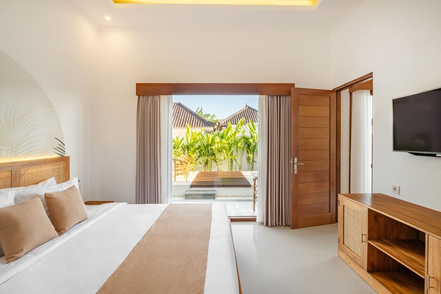 A modern bedroom with a large bed, a TV, and a view of a garden and building outside.