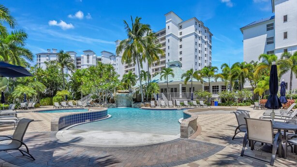 A pool area with lounge chairs and a waterfall feature, surrounded by palm trees and a multi-story building. - Windward at Regatta - Modern vacation destination! (Naples)