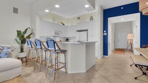 A modern kitchen with white cabinets, a central island, and bar stools. There is a dining area with a table and chairs, a potted plant, and a lamp. - Windward at Regatta - Modern vacation destination! (Naples)