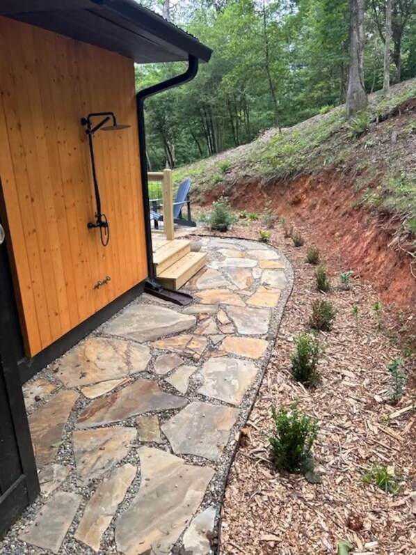 Ein Kiesweg führt zu einer Holztür mit einer Metallklinke, umgeben von einem angelegten Garten mit kleinen Pflanzen und einer Holzdar Terrasse mit einem Stuhl.
