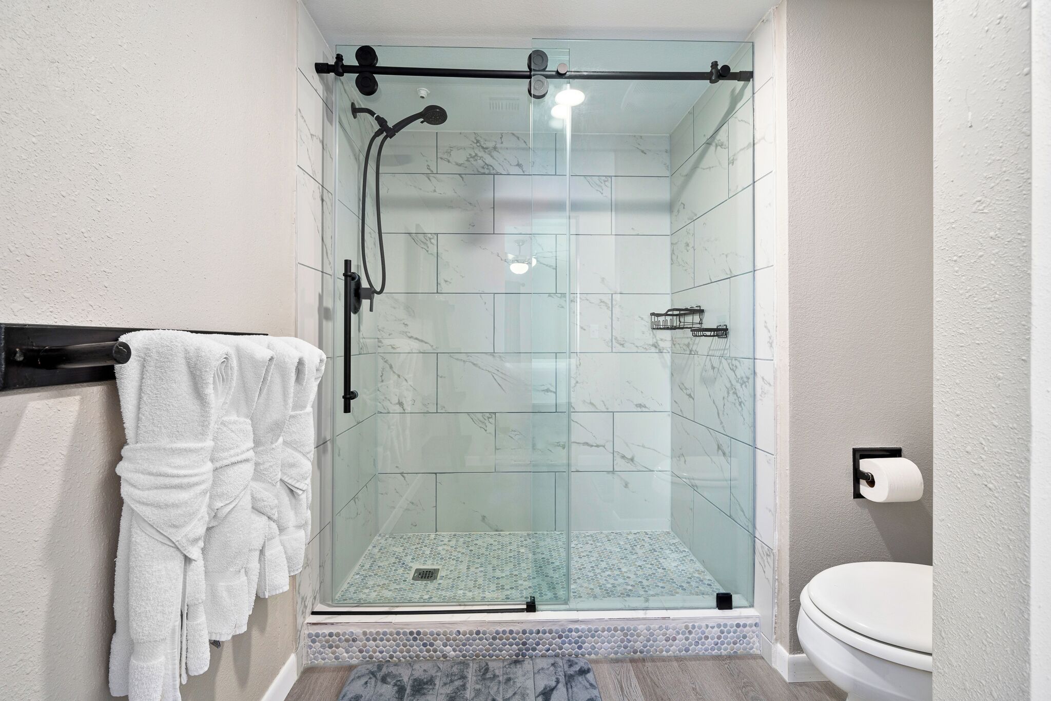 Une douche fermée par des parois de verre, avec un mur recouvert de carreaux de marbre, des toilettes et un porte-serviettes avec des serviettes.