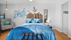 Une chambre avec un lit, un ventilateur de plafond, une tête de lit en bois, une décoration murale en forme d’ancre et un mur orné d’une enseigne sur le thème de la plage.