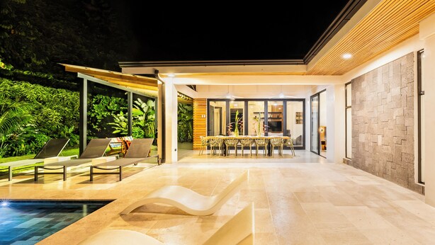 A modern outdoor pool area with lounge chairs, a dining table, and a stone wall. - Villa Bosque (La Fortuna)