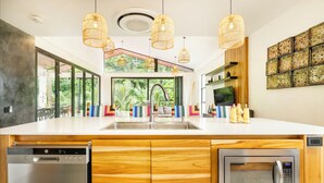 A modern kitchen with wooden cabinets, a built-in dishwasher, a microwave, and a large window overlooking greenery. - Villa Bosque (La Fortuna)
