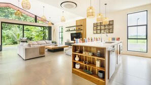 A spacious living area with a sofa, coffee table, and a wooden bookshelf. A kitchen with a sink and a large window is visible. - Villa Bosque (La Fortuna)